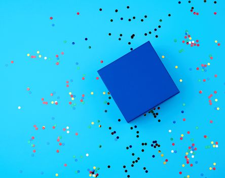 closed square empty blue gift box on a blue background, near multi-colored sequins, top view, festive backdrop