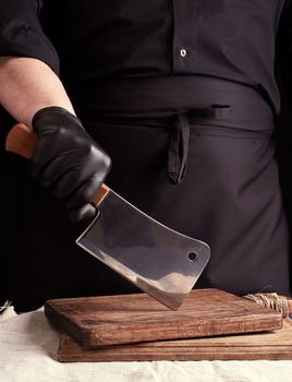 male cook in black uniform and black latex gloves holds a large sharp meat knife over a cutting board, black background