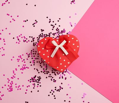 closed red heart-shaped cardboard box on a pink background with multi-colored shiny confetti, festive backdrop for birthday, Valentine's day