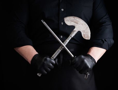 male cook in black uniform and black latex gloves holds a large sharp vintage knife for meat and vegetables and a metal sharpener for knives, black background