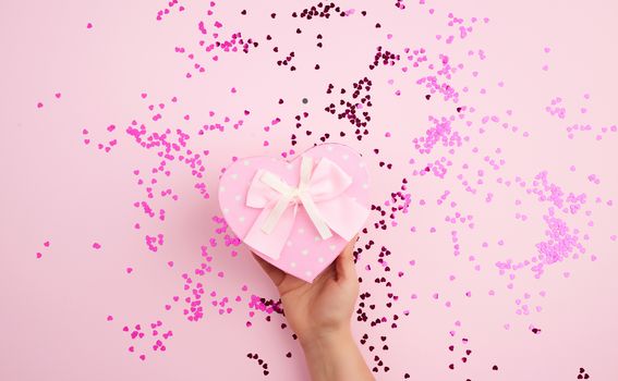 female hand holding a pink cardboard box with a bow on a pink background with multi-colored confetti, concept of giving a gift for a holiday
