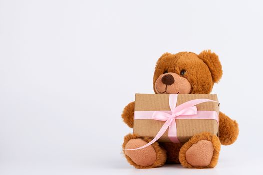 cute little brown teddy bear holds a brown box with a pink ribbon on a white background, place for a congratulatory signature