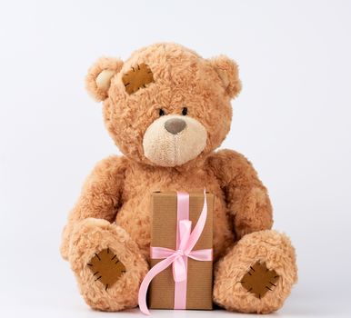 big beige teddy bear with patches hold gift in boxe wrapped in brown paper, tied with pink silk ribbon on a white background, happy birthday and valentines day