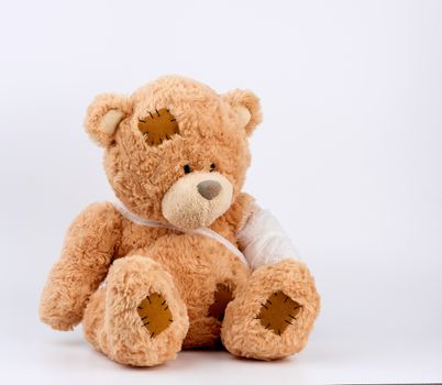 large beige teddy bear with patches sits on a white background, left paw is bandaged with a white medical bandage, concept of pediatrics, treatment of animals