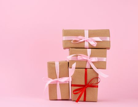 stack of gifts in boxes wrapped in brown kraft paper and tied with silk ribbon on a pink background. Festive concept