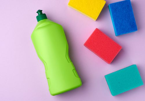 multicolored kitchen rectangular dishwashing sponges and green plastic bottle with detergent, purple background