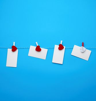 white empty paper rectangular business cards on decorative clothespins with a red heart hanging on a rope, blue background, place for an inscription