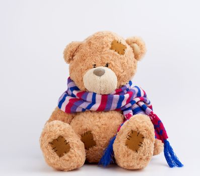 cute brown teddy bear with patches in a colored knitted scarf sitting on a white background, close up