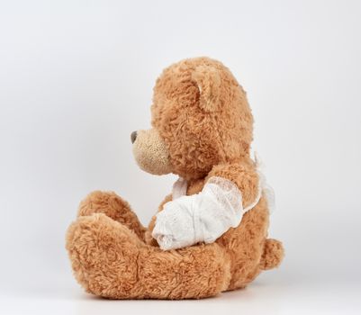 large beige teddy bear with patches sits sideways on a white background, left paw is bandaged with a white medical bandage, concept of pediatrics, treatment of animals