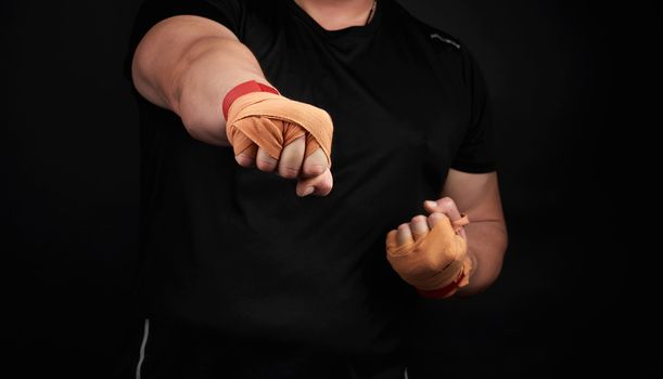 adult man in a black uniform and muscular arms stands in a sports stance, holds a sports elastic bandage on his hands, punch forward, dark background