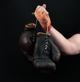 athlete in a black uniform holds very old brown boxing gloves in his hand, his hands are bandaged with an orange elastic sports bandage, black background