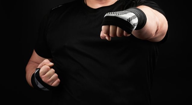 adult man in a black uniform and muscular arms stands in a sports stance, holds a sports elastic bandage on his hands, dark background