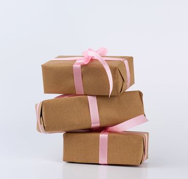 stack of gifts in boxes wrapped in brown kraft paper and tied with pink silk ribbon on a white background. Festive concept, copy space