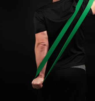 adult athlete in black uniform stands with his back and stretches a green sports elastic band, exercises for the muscles of the arms, dark background