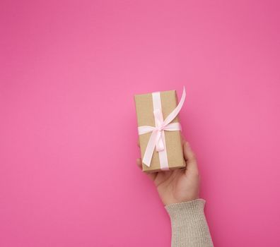 female hand holds a brown box with a pink bow on a pink background, concept of a congratulation on a happy birthday, place for text