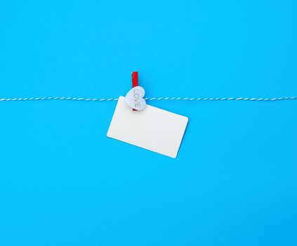 white empty paper rectangular business card on decorative clothespin with a red heart hanging on a rope, blue background, place for an inscription