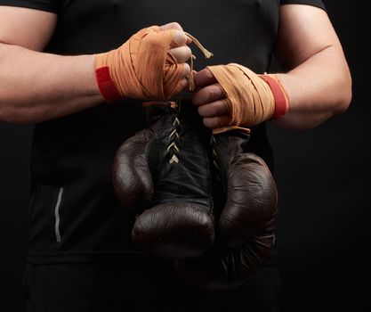 athlete in a black uniform holds very old brown boxing gloves in his hand, his hands are bandaged with an orange elastic sports bandage, black background, copy space