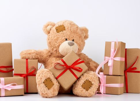 big beige teddy bear with patches, a stack of gifts in boxes wrapped in brown paper, tied with silk ribbon on a white background, happy birthday and valentines day
