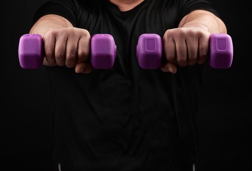 adult male athlete in a black uniform holds purple plastic blue dumbbells in hands, black background. Bodybuilding and fitness. Healthy active lifestyle