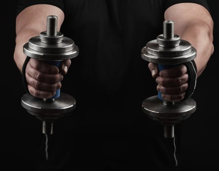 adult man in black clothes holds steel dumbbells in his hands, his muscles are tense, low key, sports background
