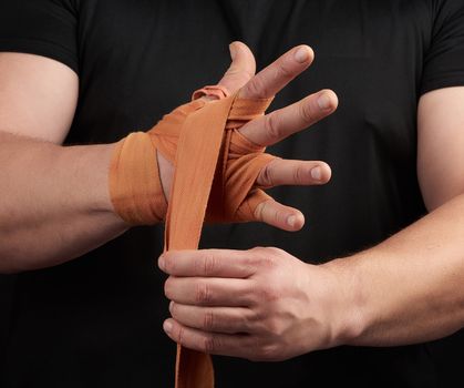 adult athlete stand in black clothes and wrap his hands in red textile elastic orange bandage before training, black background, muscular body
