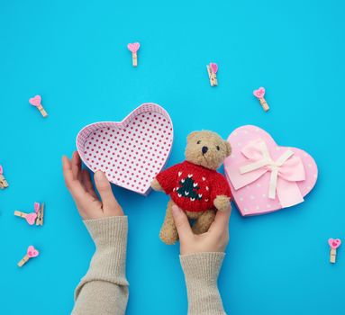 female hands holding a small brown teddy bear, on a blue background an open empty heart-shaped box, top view, holiday Valentine's Day
