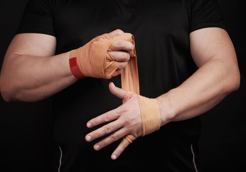 athlete stand in black clothes and wrap his hands in red textile elastic orange bandage before training, black background, muscular body