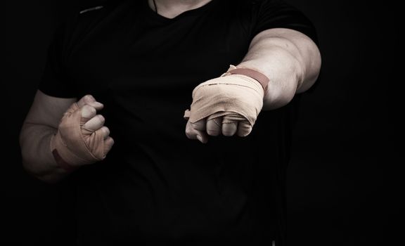 adult man in a black uniform and muscular arms stands in a sports stance, holds a sports elastic bandage on his hands, punch forward, dark background