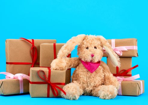 cute little brown plush rabbit and a stack of boxes wrapped in brown kraft paper and tied with silk ribbon on a blue background, concept of holiday, easter