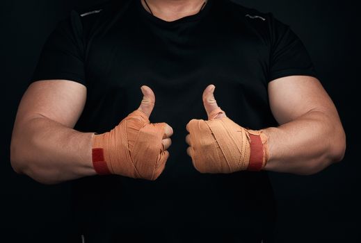 muscular athlete in black uniform shows a like symbol with two hands, palms wrapped in orange textile sports bandage, black background