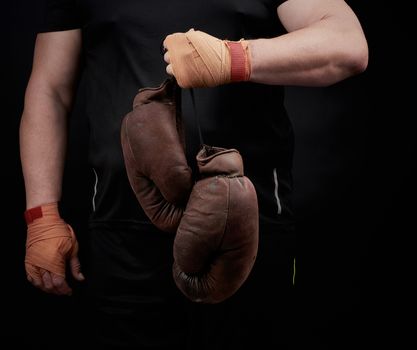 muscular athlete in a black uniform holds very old brown boxing gloves in his hand, his hands are bandaged with an orange elastic sports bandage, black background