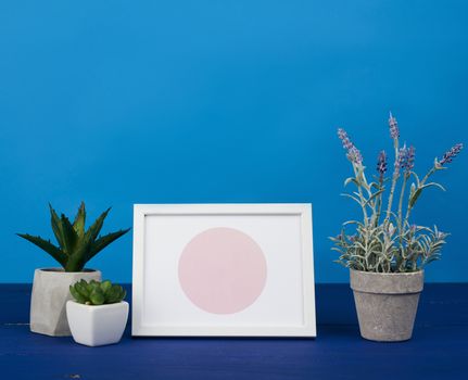 white photo frame and ceramic pot with a growing plant on a blue wooden table, minimalist style