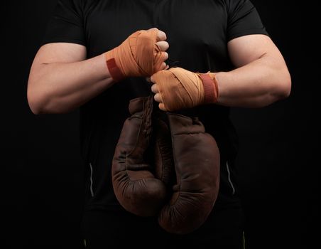 muscular athlete in a black uniform holds very old brown boxing gloves in his hand, his hands are bandaged with an orange elastic sports bandage, black background