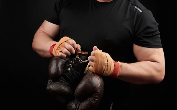 muscular athlete in a black uniform holds very old brown boxing gloves in his hand, his hands are bandaged with an orange elastic sports bandage, black background, copy space