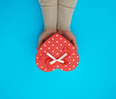 female hand holding a red cardboard box with a bow on a blue background, top view