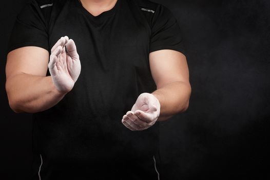 muscular athlete in a black uniform rubs his hands with white dry sports magnesia, powder scatters in different directions on a black background, copy space