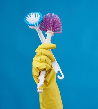 hand in a yellow rubber glove for cleaning a house holds two plastic brushes with a handle on a blue background