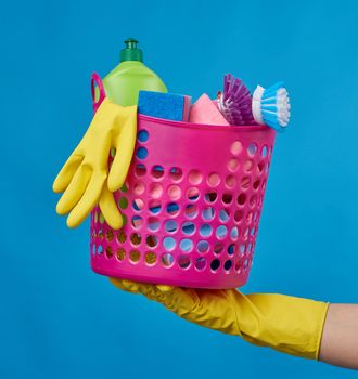 hand hold pink basket with washing sponges, rubber protective gloves, brushes and cleaning agent in a green plastic bottle on a blue background, set