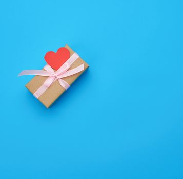 box is wrapped in brown paper and tied with a red thin silk ribbon on a blue background, red cut out paper hearts, copy space, holiday is Valentine's Day