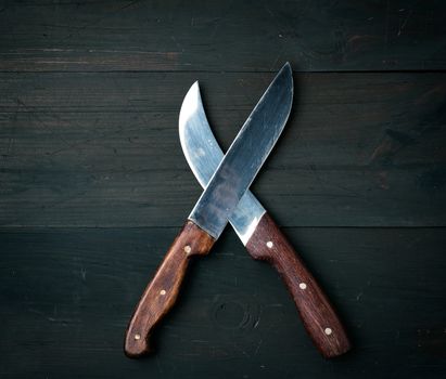 two sharp knives lie on a brown wooden surface, kitchen items are crossed, top view