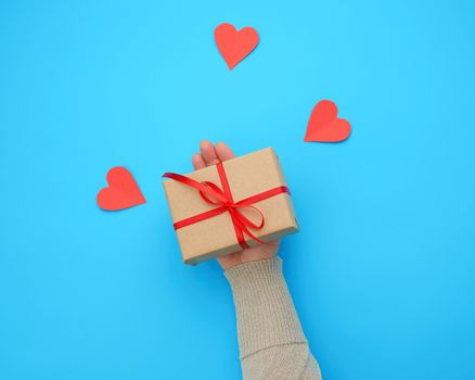 female hand holds a box wrapped in brown kraft paper and tied with a silk ribbon on a blue background, paper red hearts. Valentine's Day backdrop