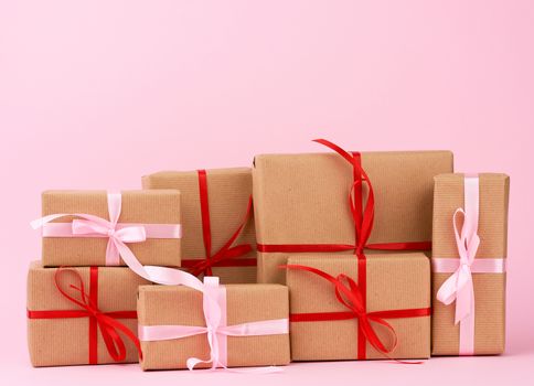 stack of gifts in boxes wrapped in brown kraft paper and tied with silk ribbon on a pink background. Festive concept, copy space