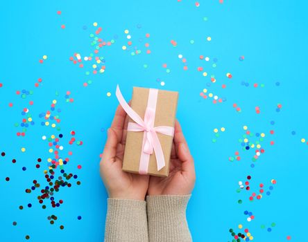 two female hands holding a brown box with a pink bow on a blue background with multi-colored confetti, concept of giving a holiday gift