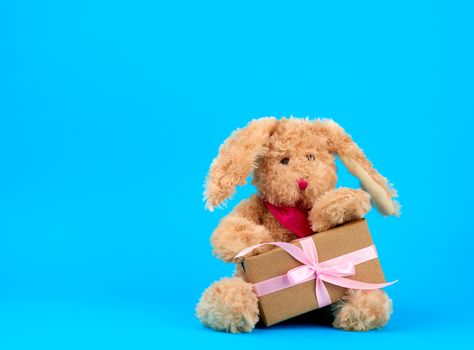 cute little brown plush rabbit  and box wrapped in brown kraft paper and tied with pink silk ribbon on a blue background, concept of holiday, easter, copy space