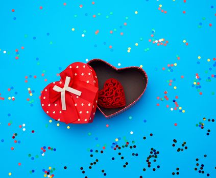 open red cardboard box in the shape of a heart on a blue background with multi-colored shiny confetti, festive backdrop for birthday, Valentine's Day