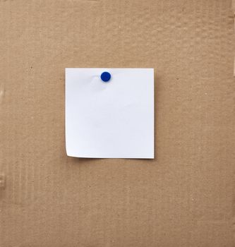 blank square white sheet of paper attached with an iron button on a brown cardboard surface. Design template.