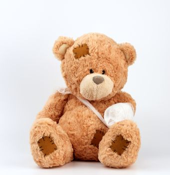 large beige teddy bear with patches sits on a white background, left paw is bandaged with a white medical bandage, concept of pediatrics, treatment of animals