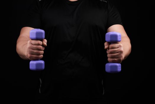 adult male athlete in black uniform holds blue plasti dumbbells in hands, black background