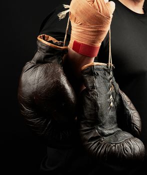 muscular athlete in a black uniform holds very old brown boxing gloves in his hand, his hands are bandaged with an orange elastic sports bandage, black background