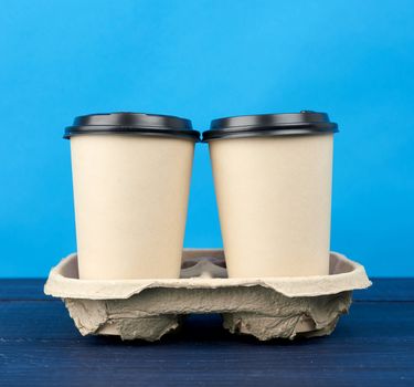 two brown paper disposable cups with a plastic lid stand in the tray on a wooden table, blue background, takeaway containers